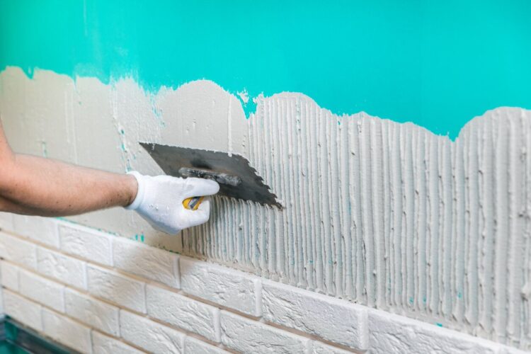 Mężczyzna w białych rękawiczkach rozprowadza klej gipsowy na niebieskiej ścianie celem przymocowania sztukaterii gipsowej.